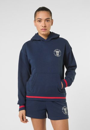 Jonge vrouw die een marineblauwe hoodie en korte broek draagt met rode strepen en witte tekst en logo op de linkerborst en dij.