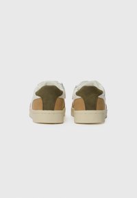Ett par vita och beige sneakers med olivgröna hälremsor, sedd bakifrån, på en enkel vit bakgrund.