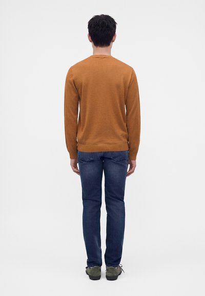 Homme debout de dos, portant un pull marron à manches longues, un jean bleu et des chaussures vertes sur un fond clair uni.