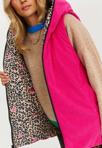 Růžový kordový vest s podšívkou s leopardím vzorem, s zipem a kapucí, nošený přes béžový svetr a modrou košili.