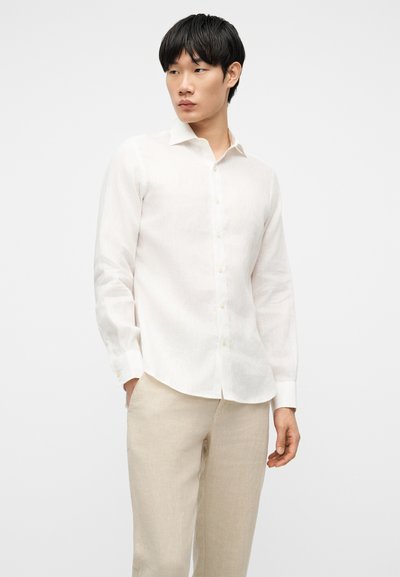 Νεαρός άνδρας που φοράει λευκό πουκάμισο με μακριά μανίκια και μπεζ παντελόνι, στέκεται με το ένα χέρι στην τσέπη, κοιτάζοντας προς τα αριστερά μπροστά σε λευκό φόντο.