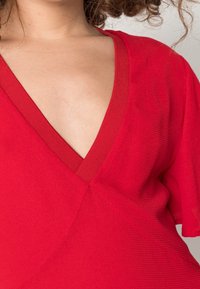 Top rojo de cuello en V con mangas cortas, que presenta una tela texturizada y un escote acanalado. El diseño incluye un detalle de costura en el centro.