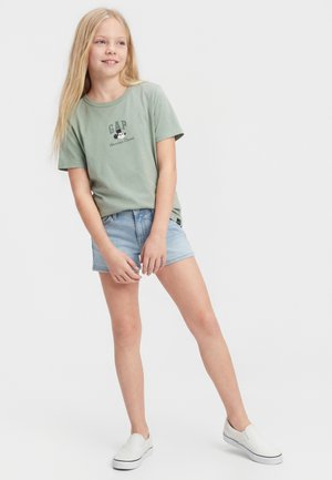 Blondína v zelenej tričku GAP s grafikou Mickeyho Mousa, rifľových kraťasoch a bielych šmykľavých topánkach, stojaca na bielom pozadí.