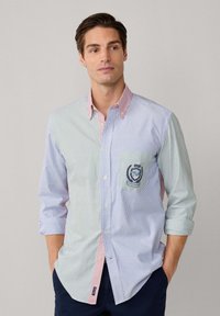 Hombre con camisa de rayas verticales multicolores de manga larga, con bolsillo en el pecho y emblema, con las manos en los bolsillos, mirando ligeramente de lado.