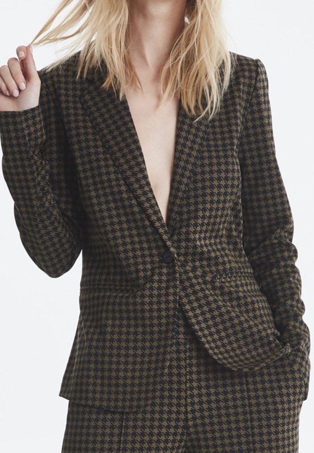 Kvinna i en brun och svart hundtandsmönstrad blazer och matchande byxor, med en hand på sitt hår och den andra i fickan.