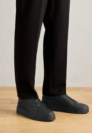 Personne vêtue d'un pantalon noir et de baskets basses noires avec lacets et fermetures éclair latérales, debout sur un sol en bois.