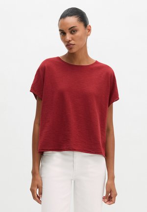 Vrouw met donker haar naar achteren gebonden, die een losse rode top met korte mouwen en een witte broek draagt, staand tegen een effen witte achtergrond.