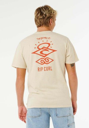 Blond man wearing beige Rip Curl T-shirt with orange geometric logo and text "The Search" on back, paired with light blue denim shorts.
