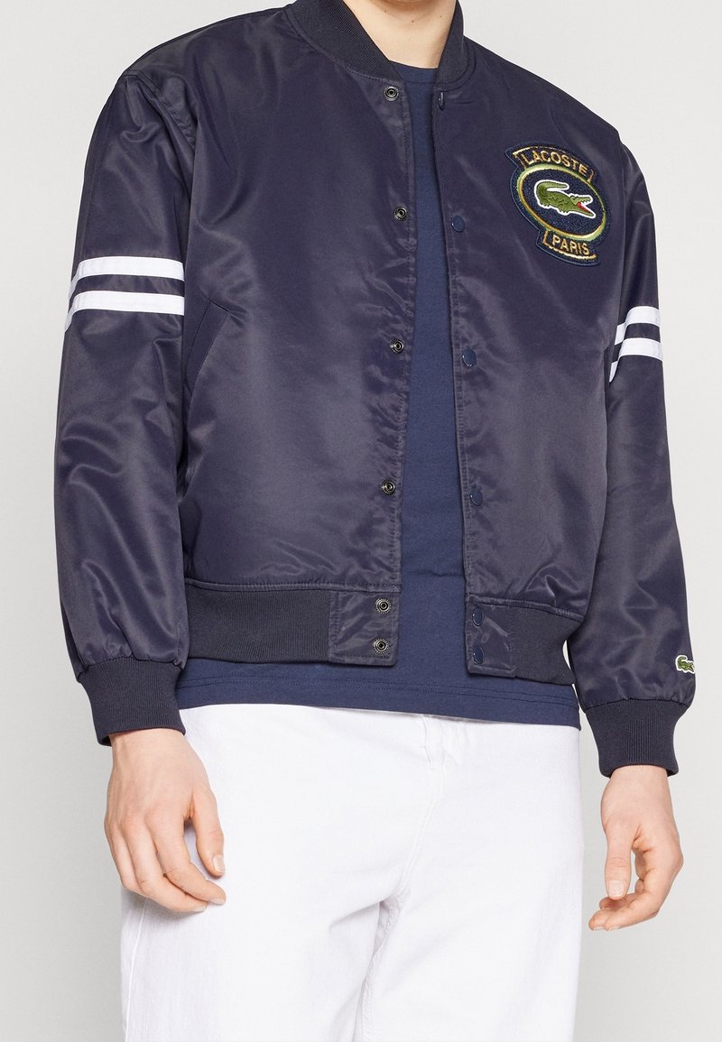 Námořnická bomber bunda vyrobená z lesklé tkaniny, se bílými pruhy na rukávech a vyšívaným logem na hrudi, s žebrovanými manžetami.