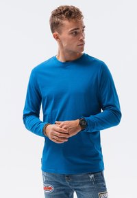 Langärmliges blaues Baumwollhemd mit rundem Ausschnitt, glatter Textur, taillierter Passform, getragen mit legeren Jeans aus Denim, die mit Flicken versehen sind.