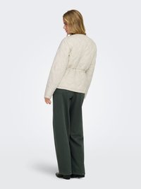 Femme aux cheveux blonds portant une veste matelassée blanche et un pantalon large vert foncé, debout et regardant vers sa gauche.