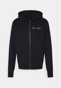 Sudadera con cremallera negra hecha de un tejido suave. Presenta un logo frontal en blanco, bolsillos laterales y puños acanalados con un acabado texturizado.