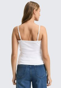 Weißes geripptes Tanktop mit verstellbaren Spaghetti-Trägern, kombiniert mit blauen Jeans mit hoher Taille und Gesäßtaschen.