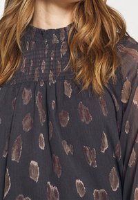Blusa scura con volant caratterizzata da una stampa leopardata in tonalità naturali, realizzata in un tessuto morbido e strutturato con una vestibilità ampia. Maniche lunghe e fluide.