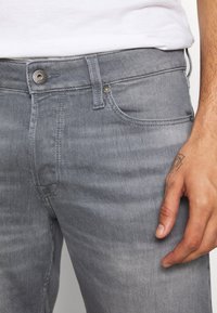 Jeans de denim gris con una cinturilla estándar, un botón plateado y bolsillos cosidos. La textura parece suave con un sutil patrón desgastado.