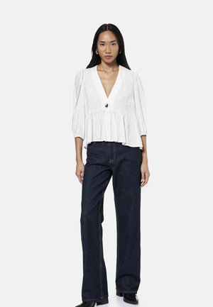 Mujer con blusa blanca de cuello en V con cintura fruncida y mangas tres cuartos, combinada con vaqueros anchos oscuros y zapatos negros.