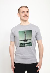 Šedé bavlněné tričko s grafikou meče na zeleném pozadí a slovy "HIGHLANDER" v tučném černém písmu pod mečem.
