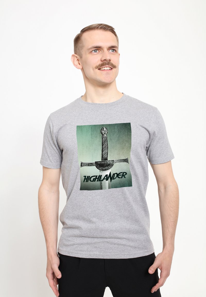 Šedé bavlněné tričko s grafikou meče na zeleném pozadí a slovy "HIGHLANDER" v tučném černém písmu pod mečem.