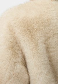 Abrigo de piel beige con una textura suave y esponjosa. La tela es densa y presenta un brillo suave, ofreciendo una apariencia rica y lujosa.