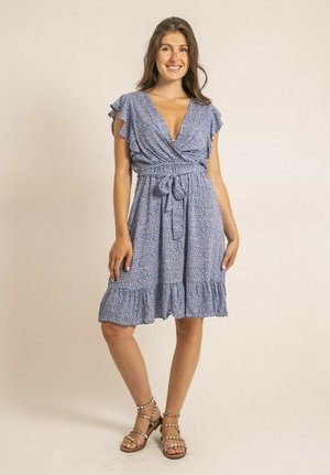 Femme souriante portant une robe bleue à fleurs longueur genou, avec des manches à volants et une ceinture à la taille, debout en sandales beiges ouvertes, sur un fond uni.