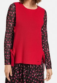 Rode gebreide mouwloze top met zijbindstrikken, gecombineerd met een zwarte blouse met lange mouwen in een kleurrijk geometrisch patroon. Textuurdetail in de stof.