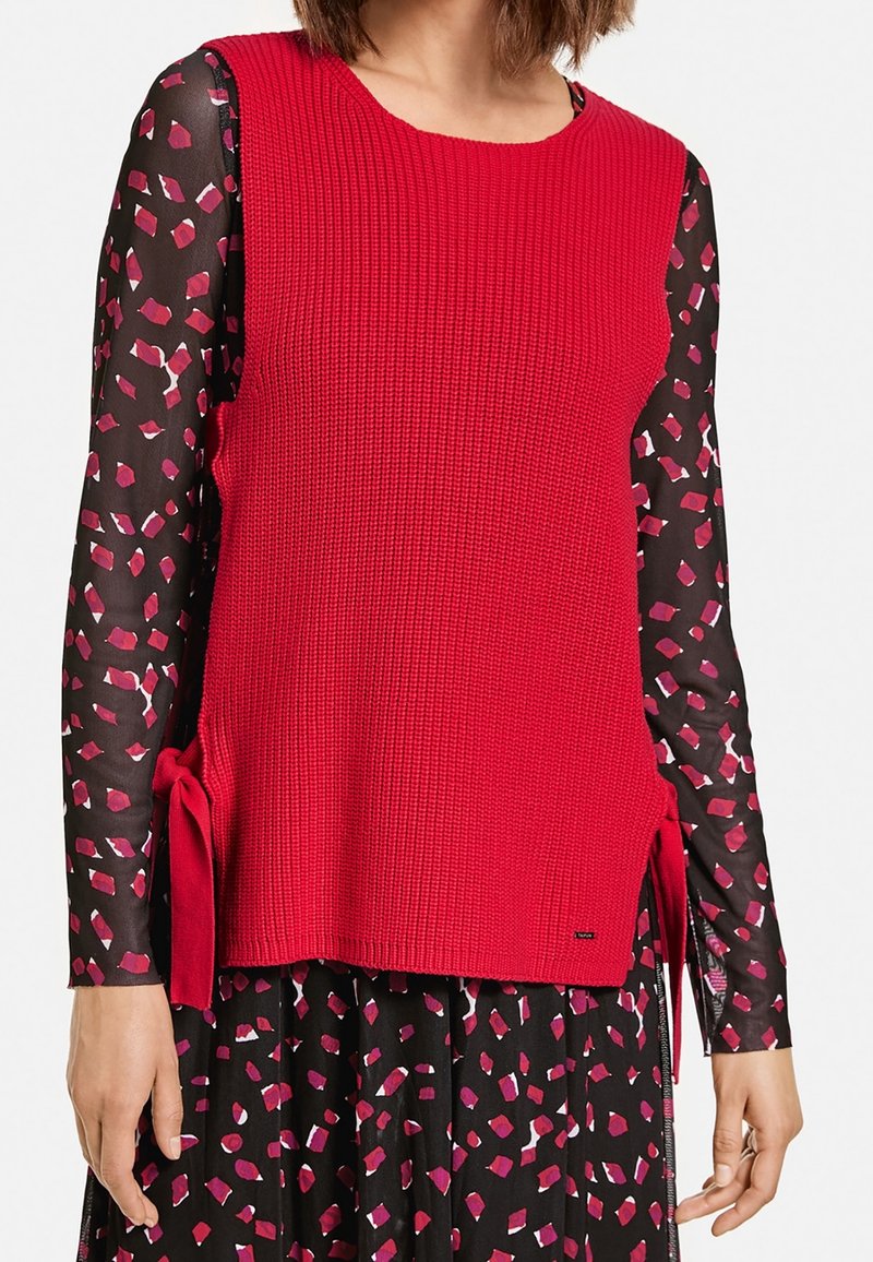 Rode gebreide mouwloze top met zijbindstrikken, gecombineerd met een zwarte blouse met lange mouwen in een kleurrijk geometrisch patroon. Textuurdetail in de stof.