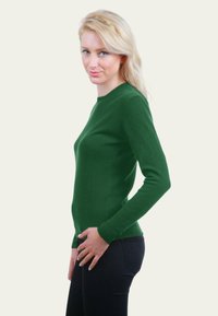 Femme blonde portant un pull ajusté à manches longues vert foncé et un pantalon noir, se tenant de profil avec une main près de la hanche.