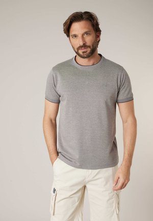 Homme barbu portant une chemise à manches courtes gris clair et un pantalon cargo blanc, debout devant un fond beige uni.
