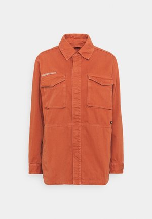 Chemise orange rouille à manches longues avec deux poches à rabat sur la poitrine et un col classique, présentée sur un fond gris clair uni.