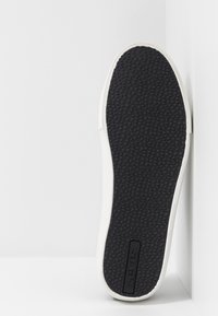 Valkoinen tennarin pohja, jossa on teksturoitu musta kumipohja. Sivussa pieni musta logo, sileät reunat ja tyylikäs muotoilu.