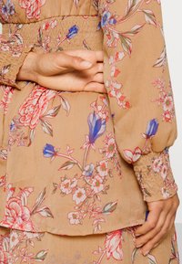 Mains reposant sur le dos d'une robe beige à fleurs avec des manches longues et des poignets smockés, présentant des motifs floraux roses, bleus et verts.