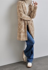 Abrigo largo de cuadros en beige y blanco con bolsillos, combinado con vaqueros azules y zapatillas blancas. El abrigo cuenta con una tela suave y texturizada.