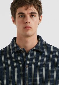 Dunkelblaues Button-Down-Hemd aus strukturiertem Stoff, mit einem weißen Gittermuster und einem Kragen. Das Hemd hat große Knöpfe und eine lässige Passform.