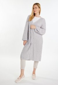 Cardigan lungo con apertura frontale in morbido maglione grigio chiaro testurizzato, caratterizzato da maniche a palloncino e una vestibilità comoda, indossato su una maglietta bianca e pantaloni beige.