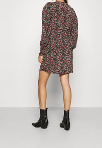 Blumenkleid in Dunkelgrün mit pinken und weißen Blumen; lange Ärmel, geraffte Schultern und ein leicht geraffter Saum, kombiniert mit schwarzen Knöchelstiefeln.
