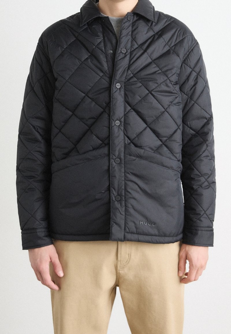 Homme portant une veste matelassée noire avec des boutons-pression et deux poches avant, associée à un pantalon beige sur un fond uni.