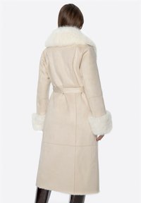 Beige suède jas met een grote witte bontkraag en bijpassende manchet. Heeft een ceintuur in de taille en een middellange snit. Gladde textuur.