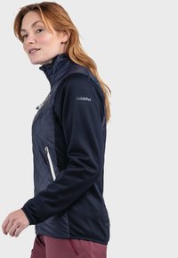 Dunkelblaue, gesteppte Softshelljacke mit Reißverschlusstaschen. Merkmale sind ein hoher Kragen und ein Logo am Oberarm. Glatte Textur und taillierte Passform.