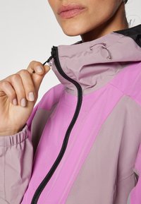 Chaqueta ligera con capucha en una mezcla de materiales morados y grises. Presenta un cierre negro, cordones ajustables y puños elásticos.