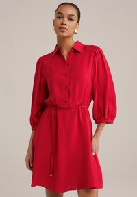 Robe rouge longueur genou avec un col boutonné, taille élastique et manches bouffantes avec une texture subtile. Lien à la taille pour un ajustement.