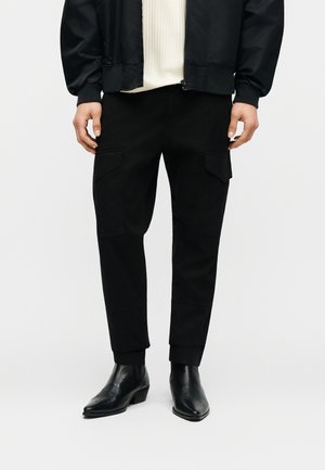 Homme portant un pantalon cargo noir avec poches à rabat, un pull en tricot blanc, une veste zippée noire et des bottines en cuir noir.