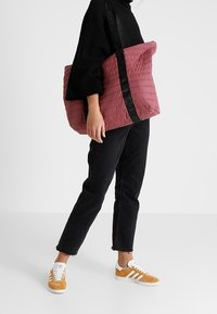 Gequilte roze totebag met zwarte schouderband, rechthoekige vorm en een gladde textuur. Draag het met zwarte jeans en tan sneakers met witte accenten.