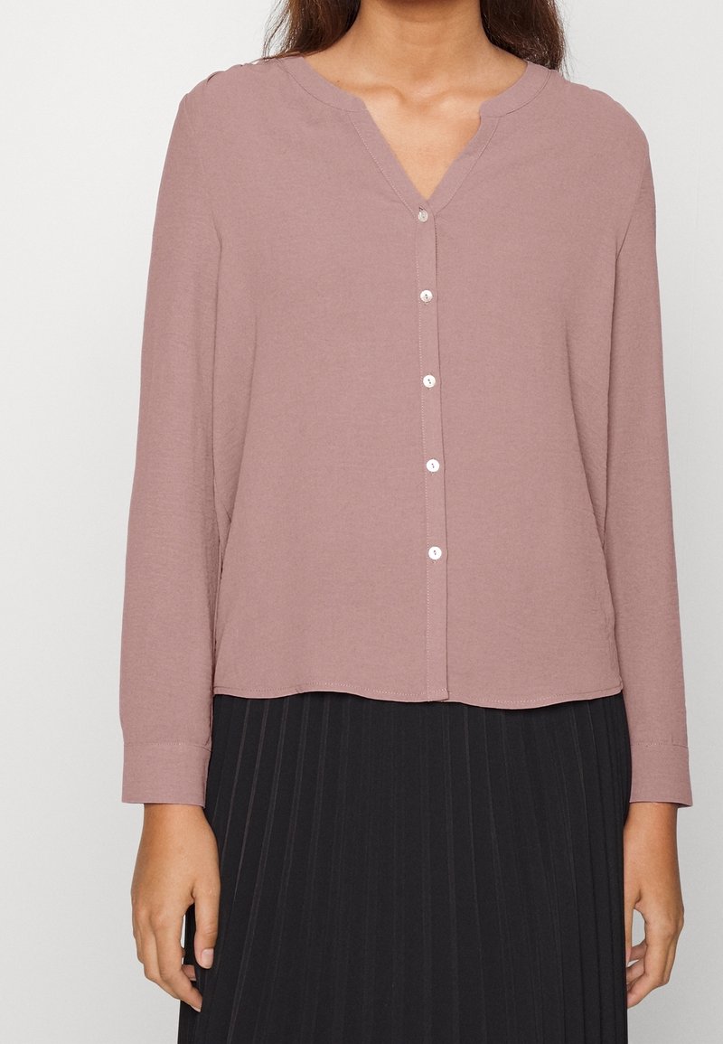 Femme portant un chemisier mauve à manches longues boutonné, rentré dans une jupe plissée noire, debout devant un fond clair uni.