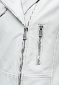 Κοντινό πλάνο λευκού δερμάτινου μπουφάν με δύο ασημένια μεταλλικά φερμουάρ, ένα διαγώνιο και ένα κάθετο στην μπροστινή πλευρά.