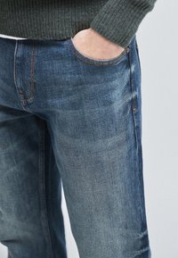 Jeans in denim blu lavaggio medio, con vestibilità classica, dettagli di cuciture visibili e una tasca frontale. Tessuto strutturato con leggera scoloritura.