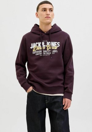 Dark purple hoodie with a front graphic, featuring a kangaroo pocket and adjustable drawstring hood. Worn with dark jeans.