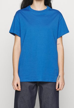 Personne portant un t-shirt bleu uni à manches courtes et un jean foncé, montrant des tatouages sur l'avant-bras gauche et le poignet droit sur un fond uni.
