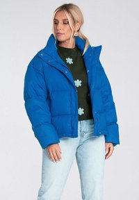 Niebieska kurtka puffer z wysokim kołnierzem, pikowanym wzorem i zatrzaskami. Noszona na zielonym swetrze w kwiatowy wzór, w połączeniu z jasnymi dżinsami.