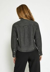 Donna con lunghi capelli castani, che indossa una giacca di jeans nera sbiadita e pantaloni neri, in piedi di fronte a uno sfondo bianco semplice.