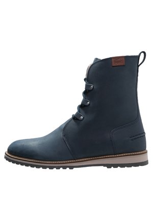 Marineblaue Leder-Stiefelette mit drei Metallösen, kontrastierender brauner Sohle und braunem Logo-Aufnäher nahe der oberen Rückseite.